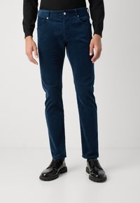 Mornarski kord tukaj s slim fit krojem in lahkotno sijajem. Ima pet žepov in kovinske gumbne poudarke. V kombinaciji s črnimi čevlji.