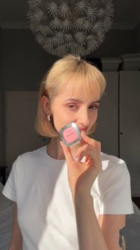 Rougekompakt i en fyrkantig silverförpackning, med ett mjukt rosa puder. Modellen håller den mot sitt ansikte och bär en enkel vit t-shirt.