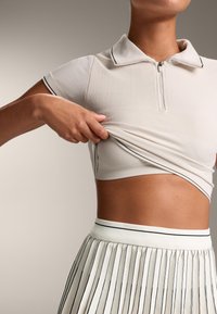 Vit polo-topp med dragkedja och krage, med textilstruktur och svarta detaljer. Matchad med en veckad kjol med vertikala ränder.