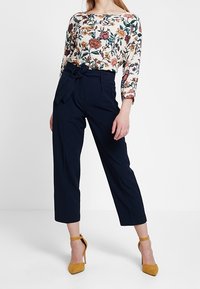 Haut à manches longues à fleurs couleur crème avec des fleurs multicolores ; pantalon taille haute bleu marine avec un détail nœud ; talons jaune à bride cheville.