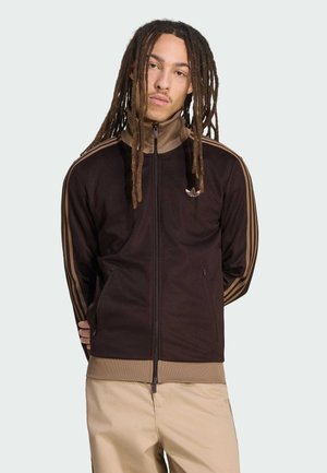 Junger Mann mit langen Dreadlocks, der eine schwarz-braune Adidas-Trainingsjacke mit Reißverschluss und beige Hosen trägt und vor einem schlichten Hintergrund steht.