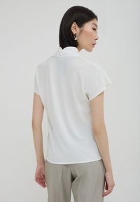 Donna con capelli corti e scuri che indossa una maglietta bianca a maniche corte e pantaloni grigio chiaro, girata di lato contro uno sfondo semplice.