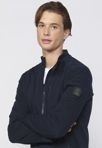 Jachetă zip-up de culoare bleumarin, cu un material texturat, guler înalt și buzunare cu fermoar. Prezintă un patch cu logo pe brațul stâng.