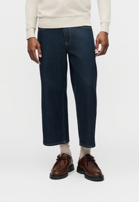 Άνδρας που φοράει κοντά σκούρα μπλε τζιν, μπεζ κάλτσες, καφέ δερμάτινα παπούτσια και μπεζ πουλόβερ με μακριά μανίκια σε λευκό φόντο.