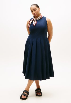 Donna in piedi con le mani dietro la schiena, indossa un abito midi senza maniche blu navy, una sciarpa al collo decorata e sandali neri su sfondo bianco.