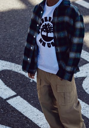 Dziecko ubrane w białą koszulkę z nadrukiem oraz otwartą zieloną koszulę w kratę i beżowe spodnie cargo stoi na asfaltowej nawierzchni z białymi oznaczeniami drogowymi.