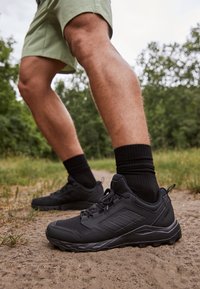 Zwarte sportschoenen van mesh en synthetische materialen, met een robuuste zool en gevoerde kraag, gedragen met zwarte sokken op een zandpad.