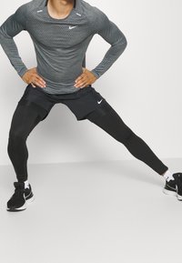 Mann in einem grauen Langarmhemd und schwarzen Shorts, der seine Beine weit auseinander dehnt, vor einem schlichten weißen Hintergrund.