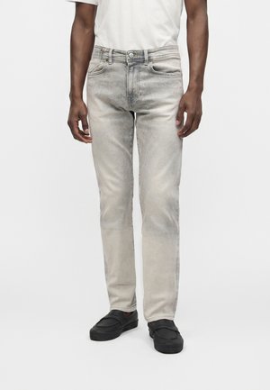 Man wearing light gray faded jeans, white shirt, and black slip-on shoes standing against a plain background.