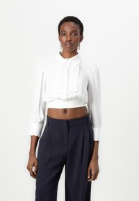 Blusa bianca corta con un colletto scalloped, maniche lunghe a sbuffo e colletto. Abbinata a pantaloni blu marine a vita alta con pieghe.