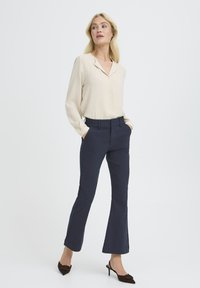 Blusa beige de manga larga con escote en V y pantalones acampanados azul marino. La modelo lleva tacones de punta marrón. Diseño simple y elegante; textura de tela suave.