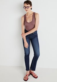Bruine geribbelde bodysuit, high-waisted donkerblauwe jeans, rode sandalen met bandjes. Model staat met arms gekruist, tegen een witte achtergrond.