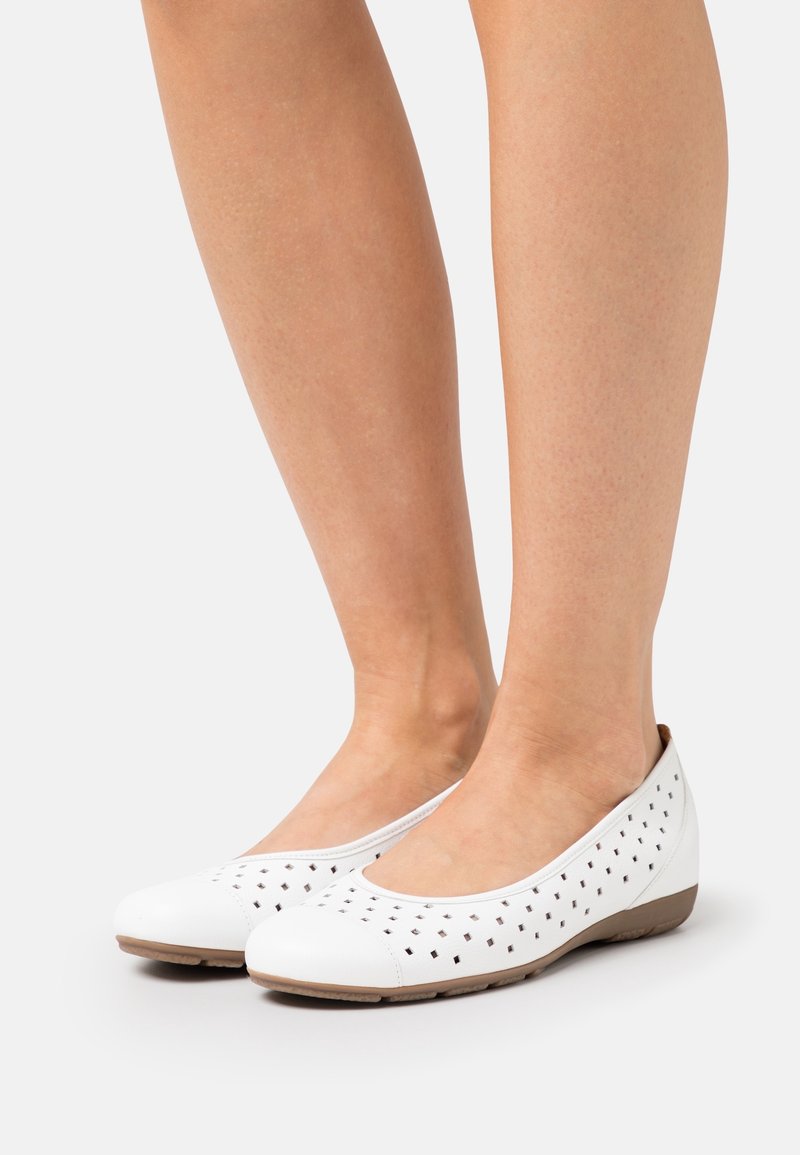 Jambes portant des chaussures plates blanches à enfiler avec des perforations carrées et des semelles marron clair sur fond uni.