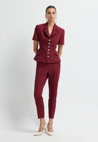 Donna in piedi su uno sfondo bianco, che indossa un blazer bordeaux a maniche corte con bottoni dorati, pantaloni coordinati, tacchi bianchi e orecchini dorati.