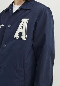Granatowa kurtka wykonana z lekkiego materiału, z dużą białą wyszywaną literą "A", zapięciem na guziki i boczną kieszenią.