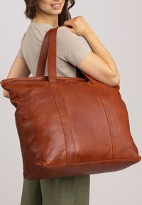 Grote bruine leren tote bag met platte handvatten, een gestructureerd oppervlak en gestikte accenten. De tas is ruim en rechthoekig van vorm.