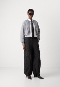 Šedá bomber bunda, bílá košile, černá kravata a oversized černé cargo kalhoty s kapsami. Černé boty s špičatou špičkou a sluneční brýle.
