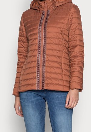 Rustfarvet quiltet jakke med dekorativ kant langs den forreste lynlås båret med blå jeans.