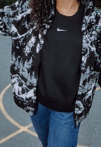 Black and white fleece jacket with abstract print, worn over a black Nike sweatshirt, paired with blue denim jeans.