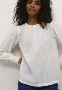 Vit blus med puffärmar i lång modell, prickig genomskinlig textur, rynkad halsringning och smockade ärmslut, ihopparad med blå jeans.