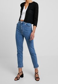 Svart kort cardigan med ett hexagonalt mönster, matchat med ljusblå högmidjade jeans och svarta remsandaler mot en neutral bakgrund.