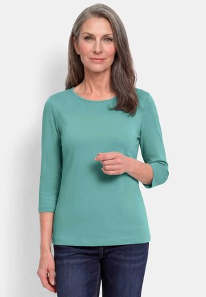 Femme aux cheveux gris mi-longs portant un haut à manches longues bleu sarcelle et un jean foncé, debout devant un fond clair et uni.
