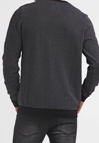 Pull en maille noir avec un motif texturé de pois, à manches ribbées et ourlet, porté sur un jean sombre. Pas de marquage visible.