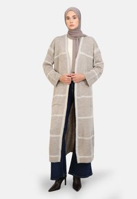 Cardigan long et ouvert beige avec un motif à carreaux blancs, avec deux poches sur le devant et une coupe décontractée, associé à un jean foncé.