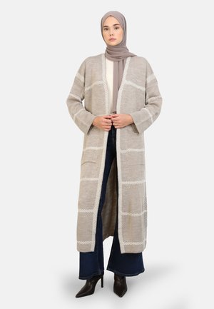 Cardigan long et ouvert beige avec un motif à carreaux blancs, avec deux poches sur le devant et une coupe décontractée, associé à un jean foncé.