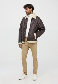 Giubbotto di pelle marrone con colletto e polsini in shearling beige, chiusura con zip e tasche laterali. Abbinato a pantaloni beige e sneakers bianche.