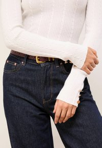 Biely rebrovaný pletený sveter s vrkočovým vzorom, s rukávmi v tvare zvončeka, doplnený tmavými džínsami a hnedým pleteným opaskom so zlatou prackou.