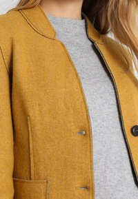 Senfgelber Wollmischjacke mit einem taillierten Kragen, grauem Pullover darunter und schwarzem Knopfdetail. Sie hat gerade Nähte als Akzente.