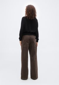 Άτομο με σγουρά μαλλιά που φοράει μαύρη μπλούζα με μακριά μανίκια και καφέ παντελόνι φαρδιάς γραμμής, όρθιο με την πλάτη στραμμένη σε ένα απλό φόντο.