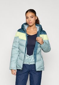 Ανοιχτό μπλε μπουφάν με κόκκινες και λαχανί λεπτομέρειες, με κουκούλα, φερμουάρ και πλευρικές τσέπες, φορεμένο πάνω από ένα σκούρο πουκάμισο.