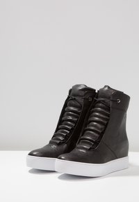 Černé kožené wedge tenisky s texturovaným, skladaným předním dílem, zaoblenou špičkou a bílou gumovou podrážkou. Vybavené kovovými očky a šněrovacím designem.