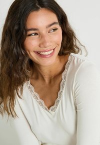 Top de manga larga en color beige claro con escote en V y delicado adorno de encaje. Textura suave con un ajuste ceñido y un diseño minimalista.