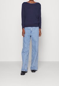 Personne portant un haut manches longues bleu marine, un jean large bleu clair et des chaussures noires à bout carré, debout contre un mur blanc uni.