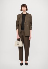 Brun skræddersyet blazer med dobbelte knapper, sort tekstureret top, løse brune bukser og en lille cremefarvet håndtaske med logo. Sorte flade sko.
