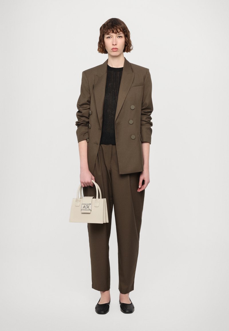 Brun skræddersyet blazer med dobbelte knapper, sort tekstureret top, løse brune bukser og en lille cremefarvet håndtaske med logo. Sorte flade sko.