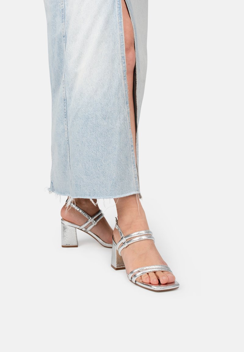Silberne Sandalen mit Blockabsatz und zwei Riemen über dem Fuß. Glänzende, metallische Oberfläche und rechteckige Zehenform. Getragen mit einem Jeansrock.
