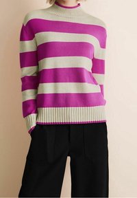 En person som bär en krämfärgad och ljusrosa randig tröja med ribbade ärmslut och nederkant, i kombination med svarta skräddarsydda byxor.