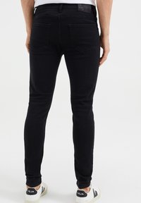 Černé slim-fit džíny vyrobené z denimu, s zadními kapsami a štítkem na pásu, spárované s bílými teniskami a černými akcenty.