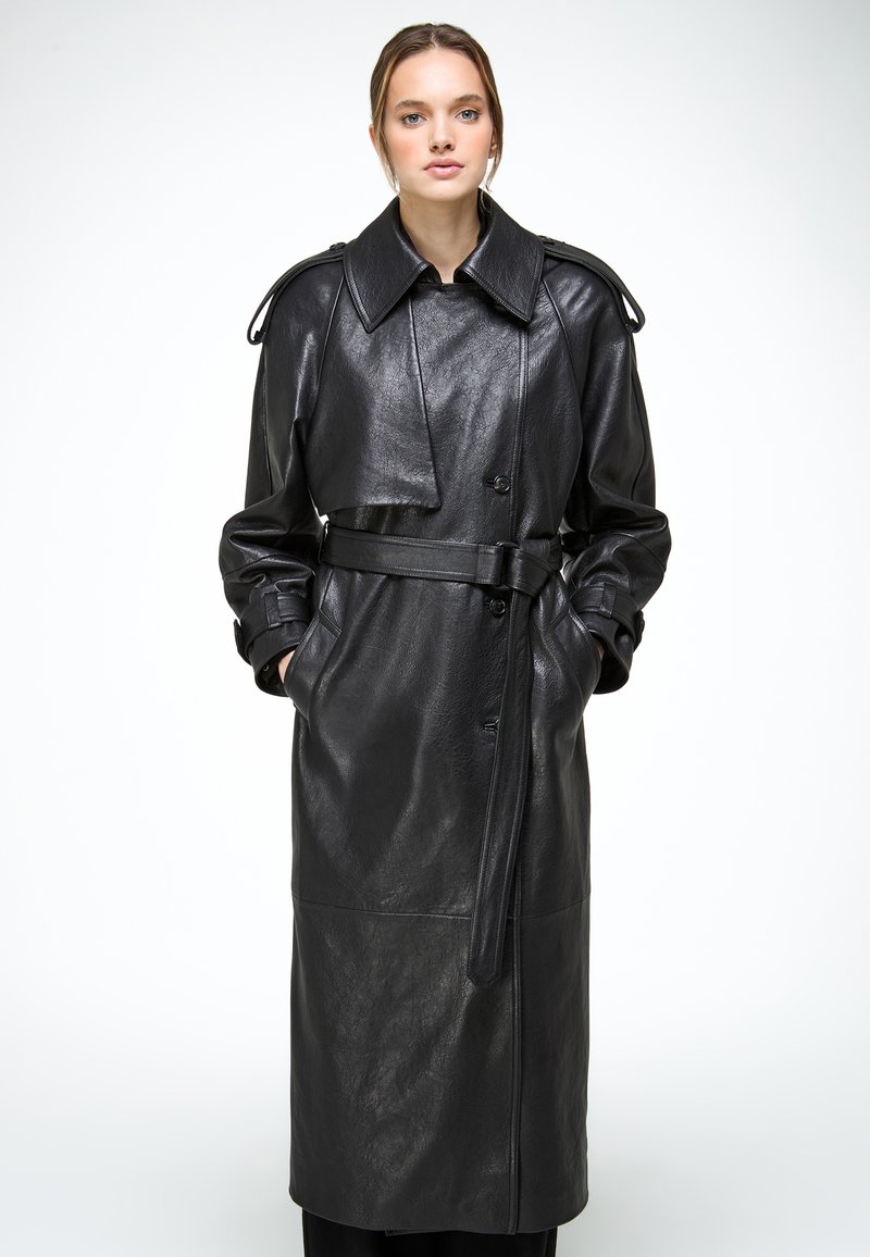 Vrouw die een lange zwarte leren trenchcoat draagt met een ceintuur in de taille, een brede kraag en handen in de zakken, staand tegen een witte achtergrond.