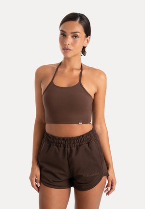 Młoda kobieta o ciemnych włosach nosi brązowy bezrękawnik typu crop top oraz pasujące krótkie spodenki z elastycznym pasem na jednolitym jasnym tle.