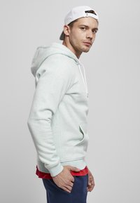 Felpa con cappuccio verde menta chiaro, dotata di tasca frontale, cordini bianchi e polsini a coste. Indossata sopra una maglietta rossa, abbinata a pantaloni della tuta blu navy.