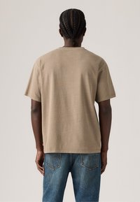 Person med flettehår iført en enkel beige kortermet T-skjorte og blå jeans, stående med ryggen til mot en nøytral bakgrunn.