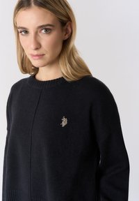 Maglione nero lavorato a maglia con scollo rotondo a coste e maniche lunghe. Presenta un piccolo logo dorato ricamato sul petto. Texture morbida.