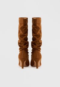 Bottes en daim beige montantes au design décontracté, avec des pointes effilées et des talons de 5 cm. La texture lisse et les accents plissés rehaussent la structure.