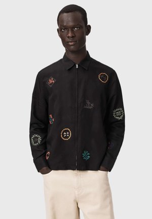 Mann trägt eine schwarze Jacke mit Reißverschluss und bunten bestickten Aufnähern sowie beige Hosen, steht vor einem schlichten hellen Hintergrund.