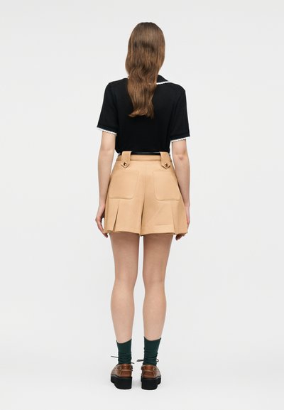 Γυναίκα που φοράει μαύρο τοπ με κοντά μανίκια και μπεζ σορτς ψηλής μέσης με μεγάλες πίσω τσέπες, πράσινες κάλτσες και καφέ παπούτσια, με την πλάτη γυρισμένη.